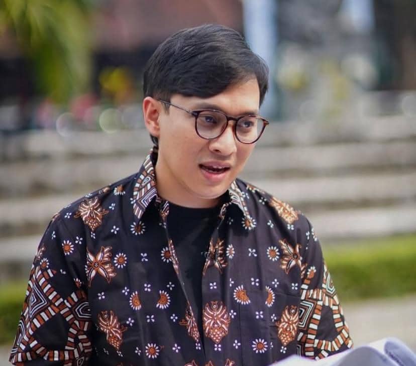Seorang pria berkacamata mengenakan kemeja batik berwarna gelap dengan motif cokelat dan putih sedang berbicara di luar ruangan.
