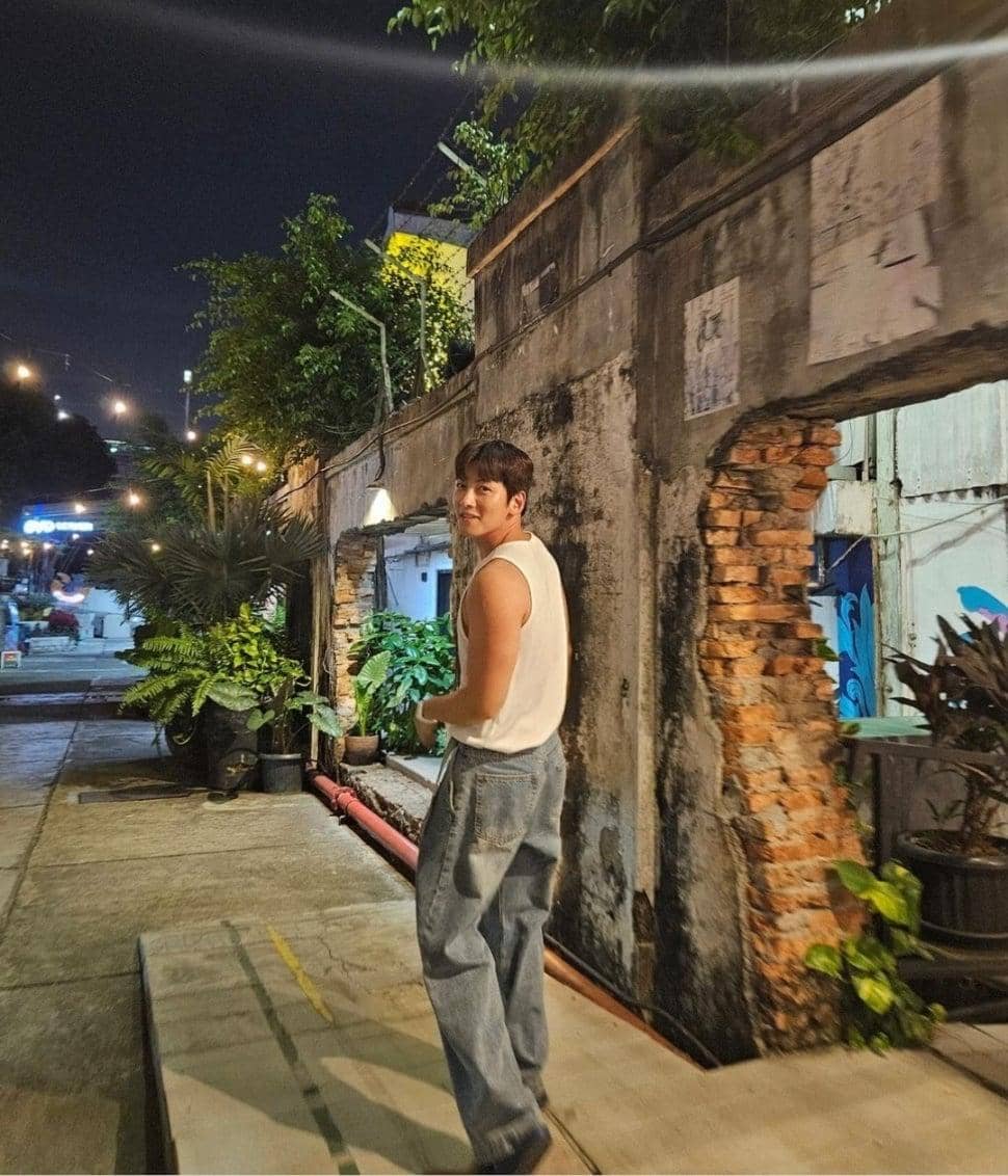 Seorang pria mengenakan kaus tanpa lengan putih dan celana jeans berjalan di jalan sempit dengan dinding bata tua pada malam hari.