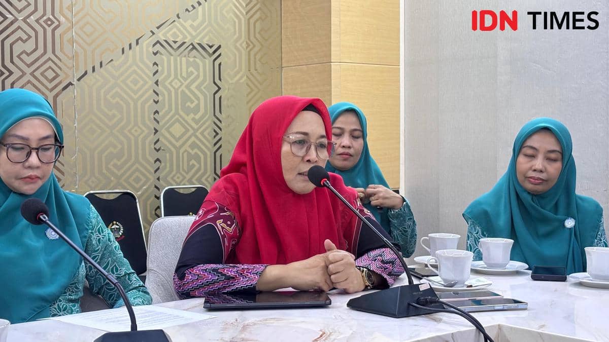 Seorang perempuan berhijab merah berbicara di depan mikrofon saat rapat bersama beberapa perempuan berhijab hijau di sebuah ruangan.