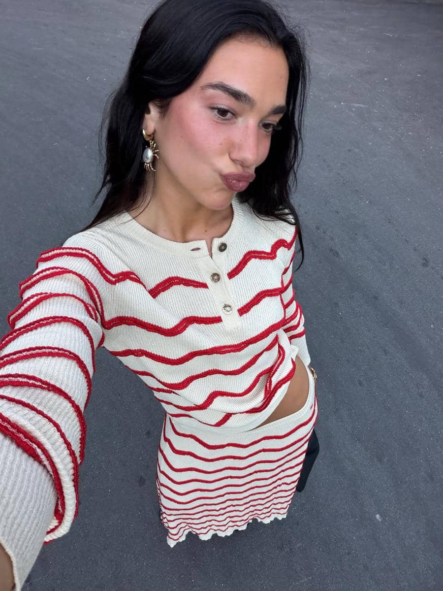 Seorang perempuan berpose selfie mengenakan atasan dan rok bermotif garis merah putih di area jalan beraspal.