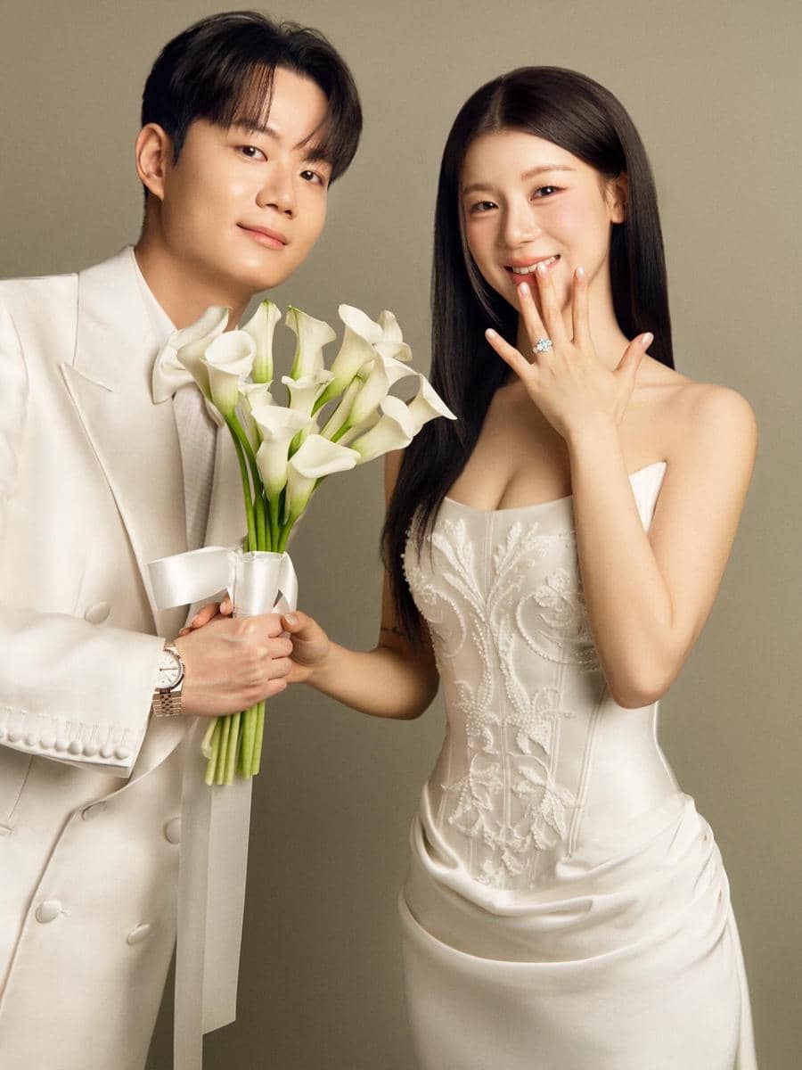 Sepasang calon pengantin mengenakan busana putih elegan, pria memegang buket bunga calla lily sementara wanita memperlihatkan cincin di jarinya.
