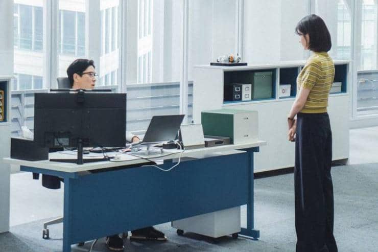Dua orang berada di kantor modern, satu duduk di meja kerja dengan komputer dan satu berdiri di depannya dalam suasana serius.
