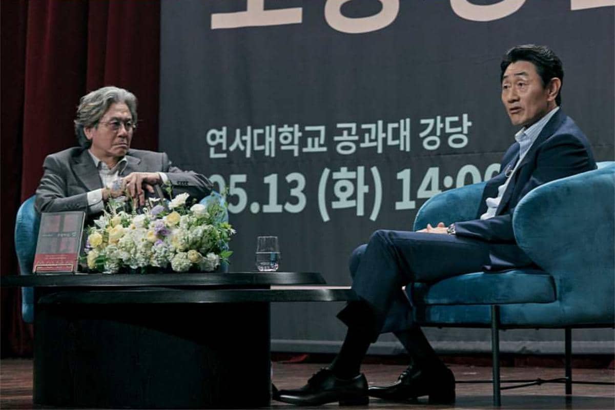 Dua pria duduk di atas panggung saat acara diskusi, dengan meja berisi bunga dan latar bertuliskan teks Korea di belakang mereka.