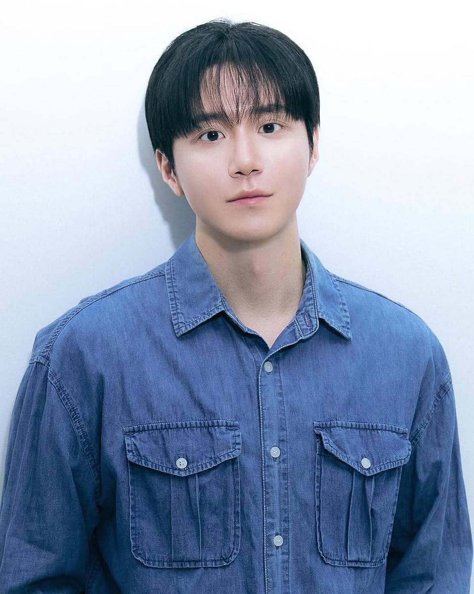 Seorang pria berambut hitam mengenakan kemeja denim biru berdiri di depan dinding putih dengan ekspresi tenang.