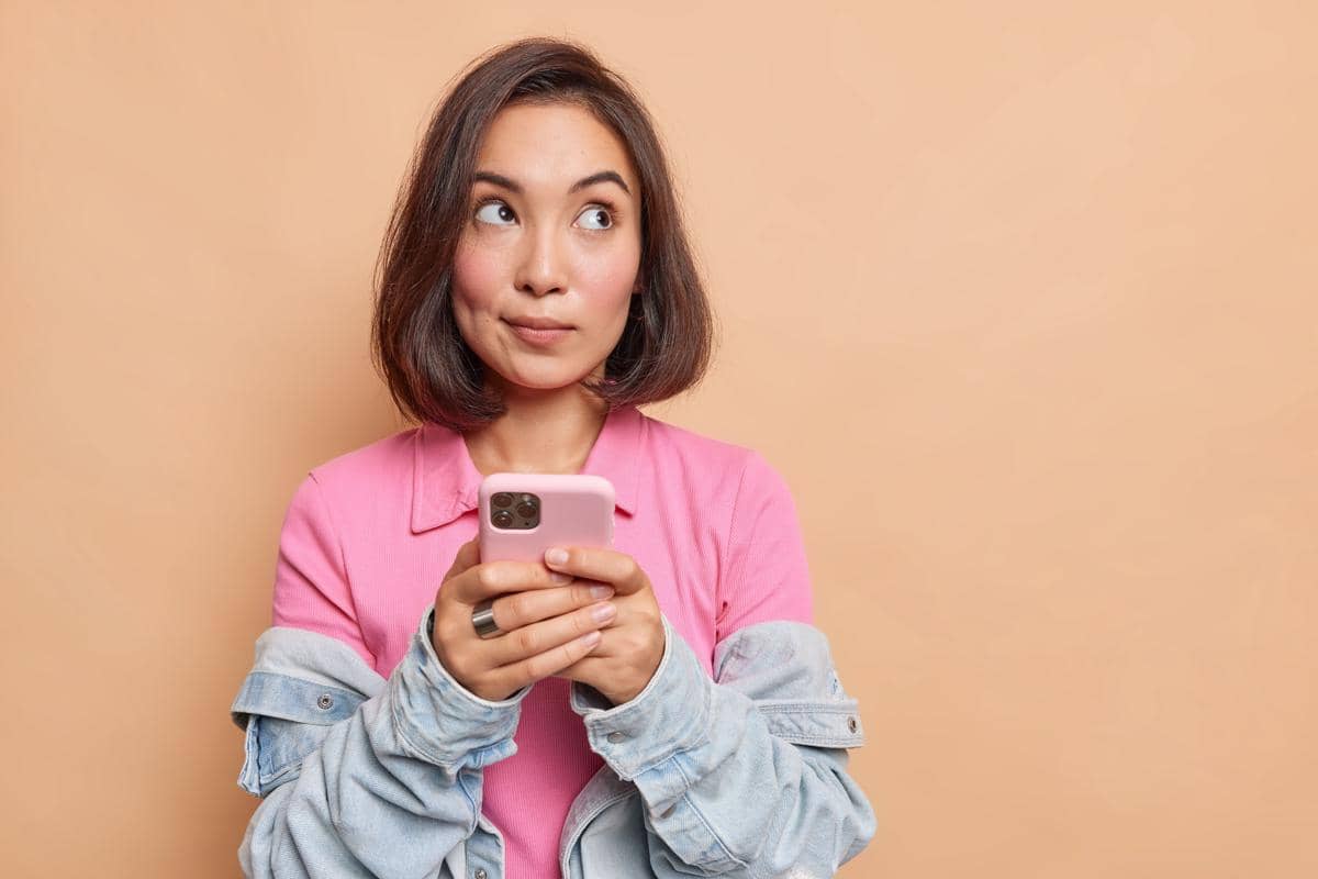 Perempuan berambut pendek mengenakan kemeja pink dan jaket denim memegang ponsel sambil menatap ke samping dengan latar oranye lembut.