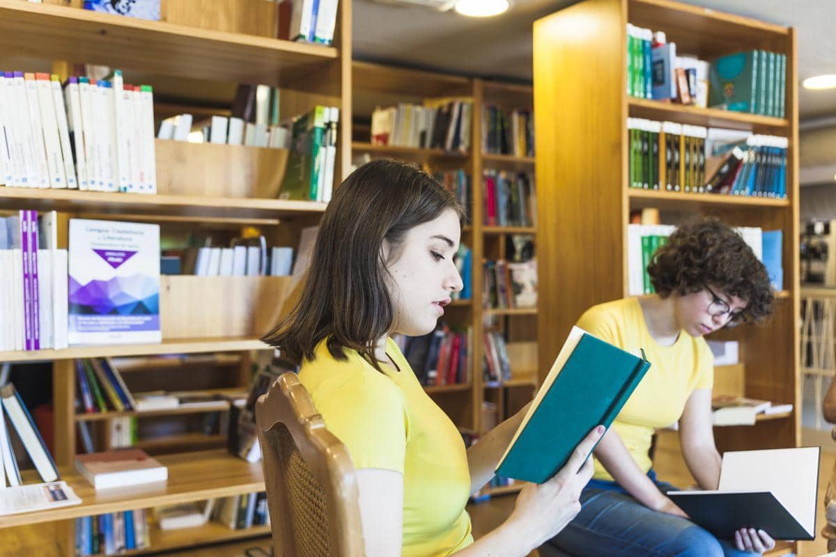 Dua orang sedang membaca buku di perpustakaan dengan rak kayu berisi berbagai koleksi buku di latar belakang.