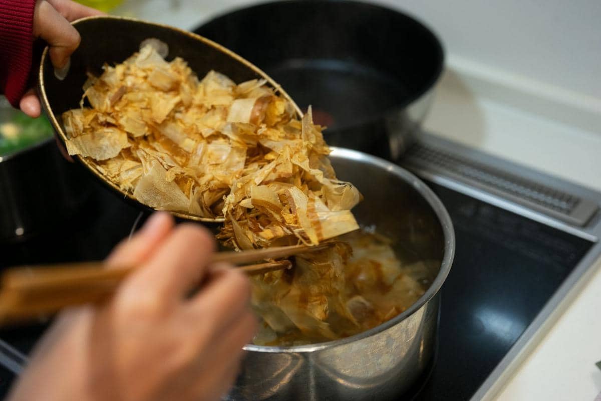 Seseorang menambahkan serpihan katsuobushi ke dalam panci berisi air panas di atas kompor untuk membuat kaldu katsuo dashi.