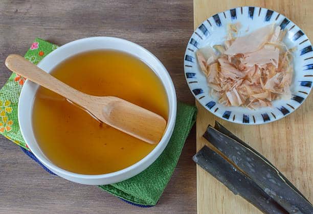 Semangkuk awase dashi dengan sendok kayu di atas kain hijau, disertai katsuobushi dan potongan kombu di atas papan kayu.