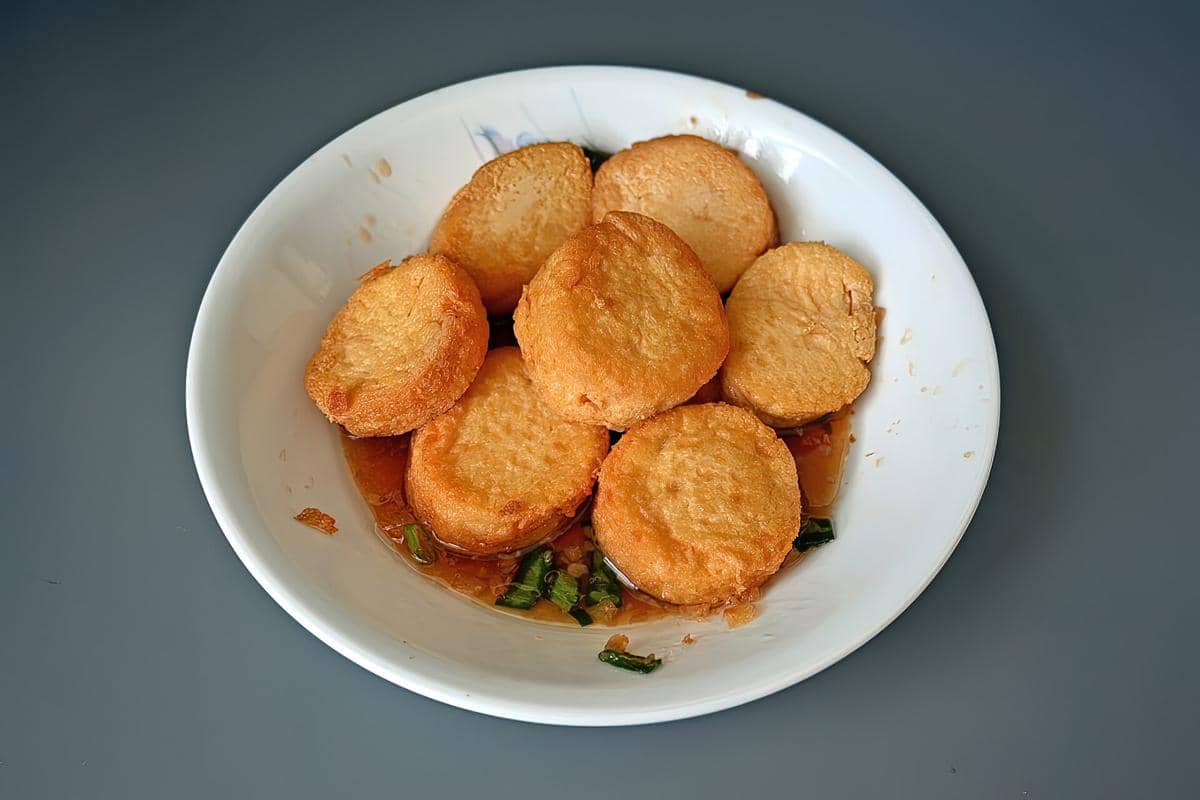 Beberapa potong egg tofu goreng berwarna keemasan disajikan di atas piring putih dengan saus kecokelatan dan potongan daun bawang.