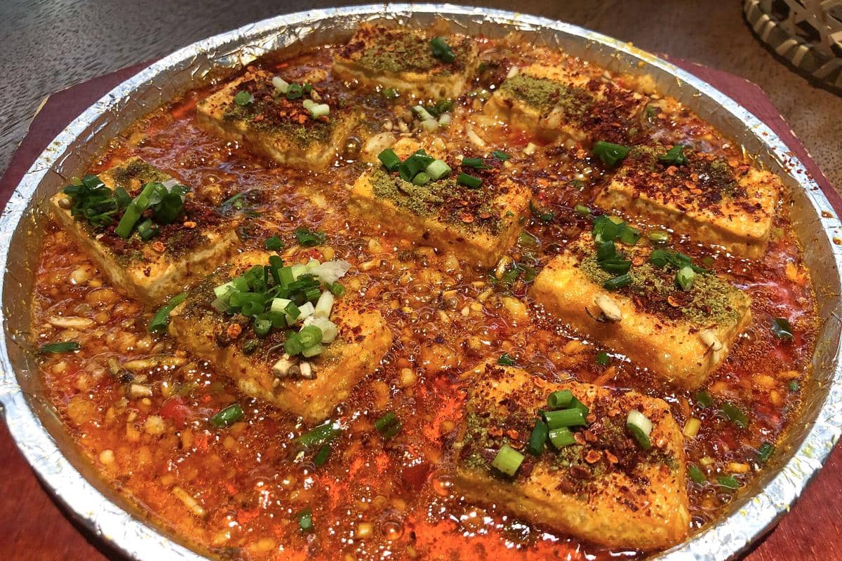 Potongan tahu baojiang disajikan dalam saus pedas berwarna merah dengan taburan cabai bubuk, daun bawang, dan bumbu rempah di atasnya.