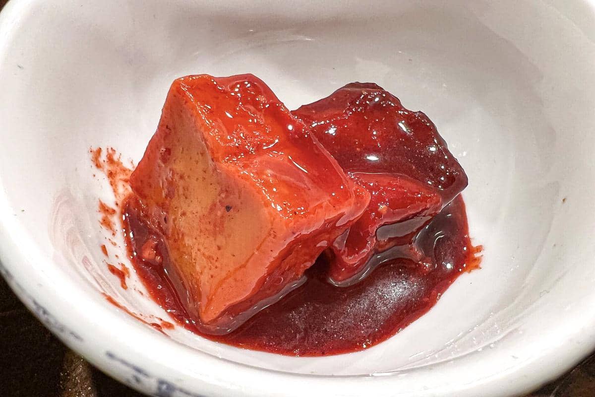 Potongan tahu fermentasi berwarna merah mengilap disajikan dalam mangkuk putih dengan saus kental berwarna merah tua.