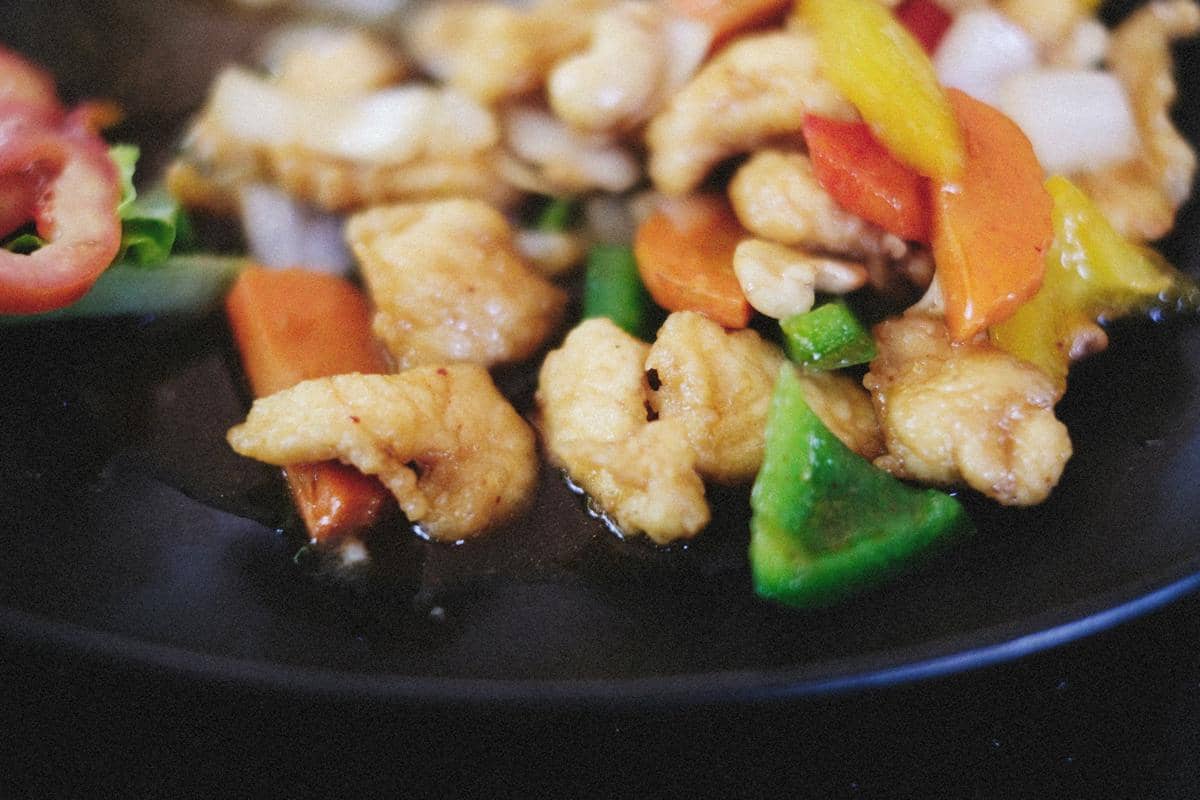 Potongan ayam velvet dimasak dengan paprika hijau, merah, dan kuning serta wortel di atas wajan hitam.