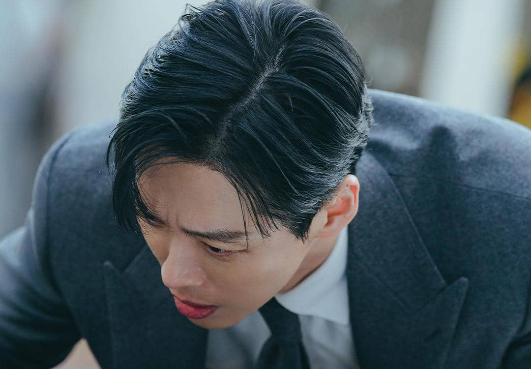 Seorang pria berjas abu-abu dengan ekspresi serius menunduk dalam adegan drama Korea The Husband.