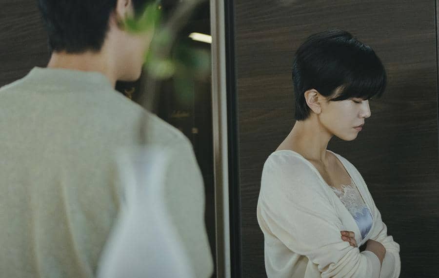 Seorang wanita berambut pendek berdiri dengan tangan terlipat dan ekspresi sedih di depan seorang pria dalam adegan drama Korea The Husband.