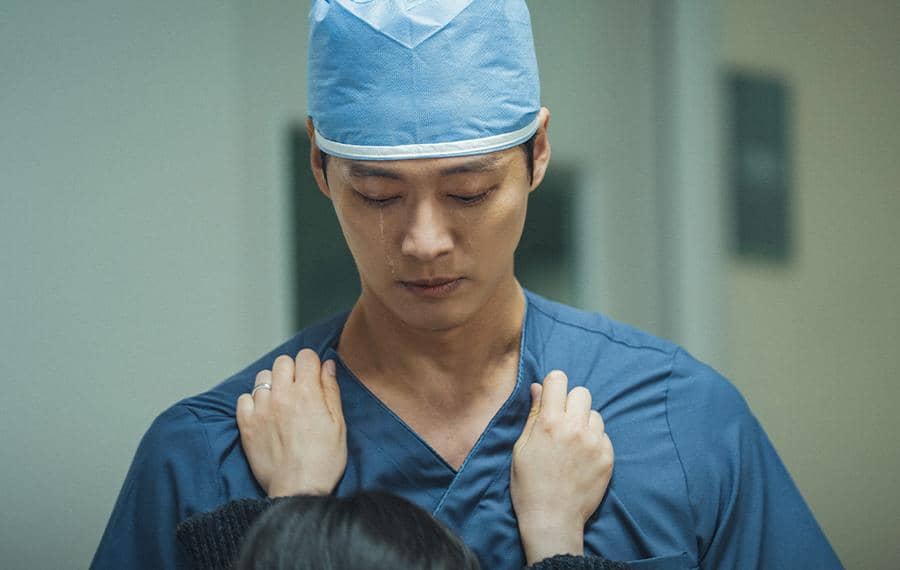Seorang pria mengenakan seragam dokter bedah dan topi medis menunduk dengan ekspresi sedih, sementara seseorang memegang bahunya.