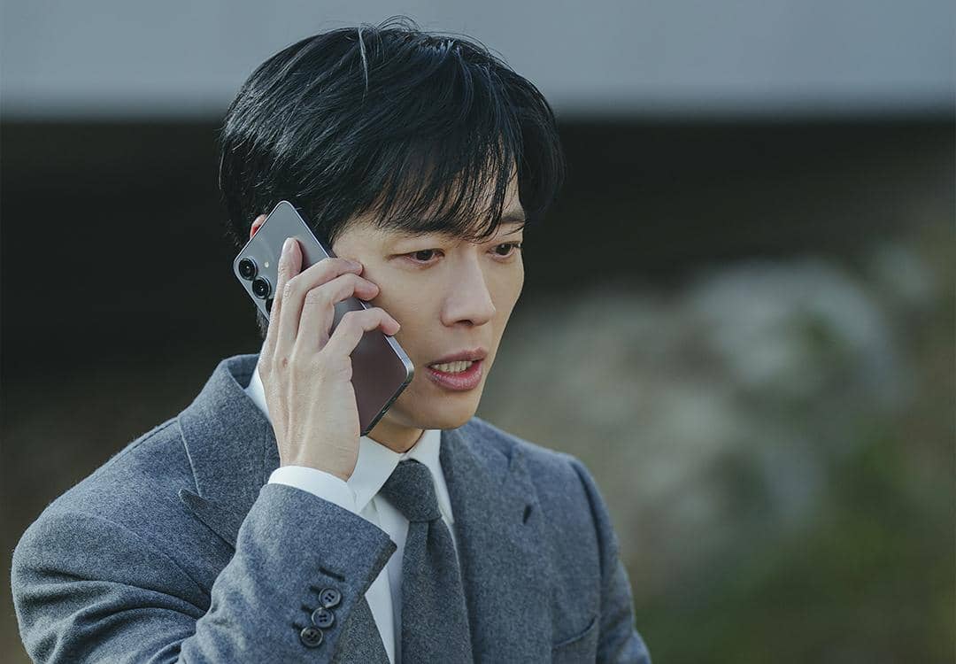 Seorang pria mengenakan setelan jas abu-abu sedang berbicara di telepon dengan ekspresi serius dalam adegan drama Korea The Husband.