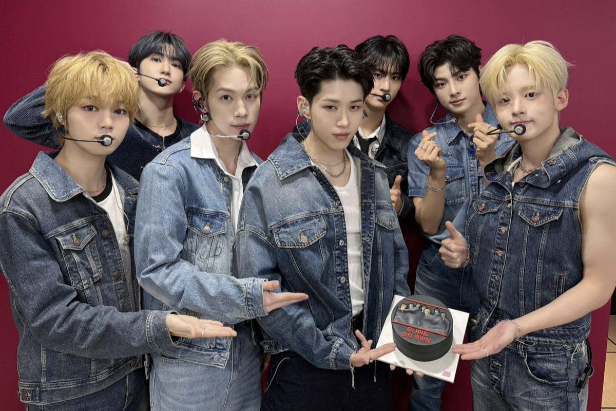 Delapan anggota grup musik pria berpose bersama mengenakan pakaian denim biru di depan latar merah sambil memegang kue perayaan.