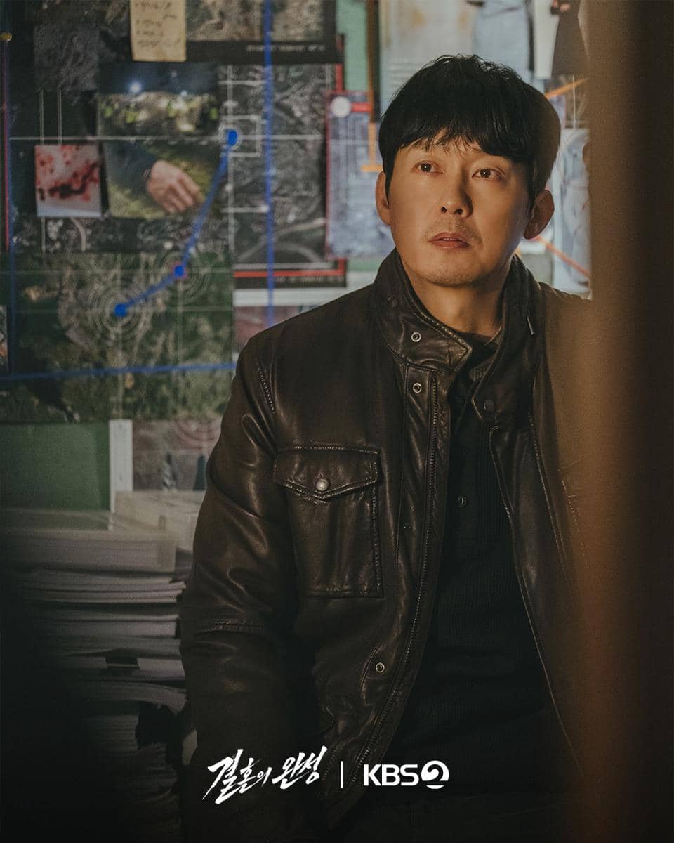 Seorang pria mengenakan jaket kulit cokelat duduk di ruangan dengan papan penuh foto dan peta di belakangnya dari drama The Husband.