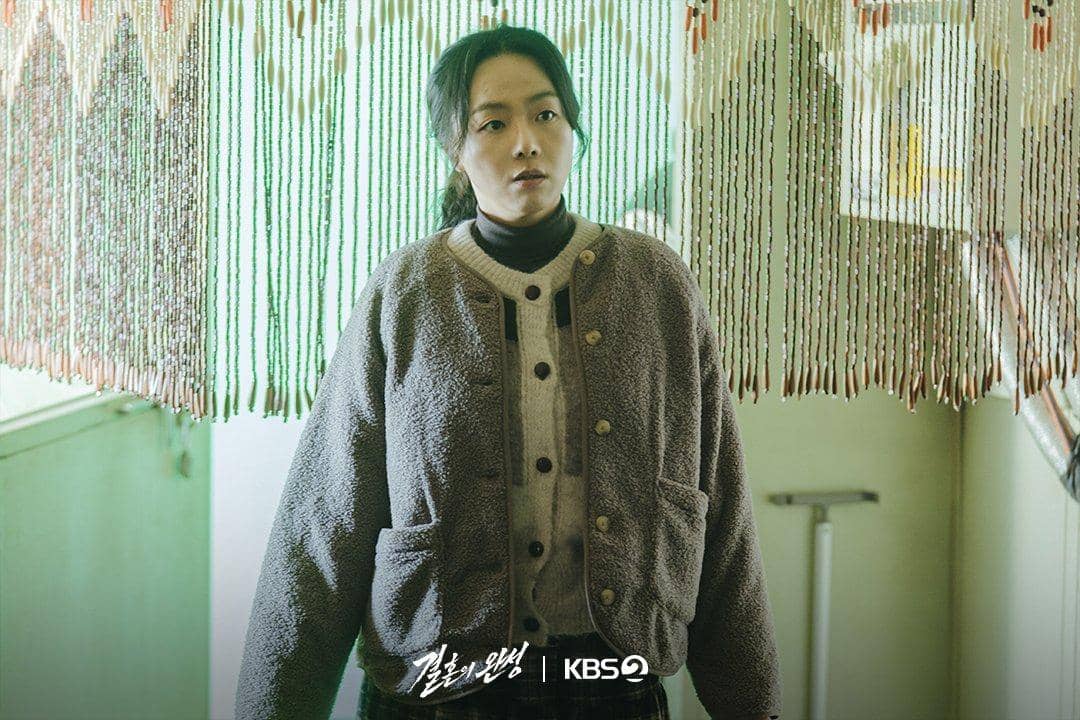 Seorang wanita berdiri di depan tirai manik-manik berwarna hijau dalam adegan drama The Husband yang ditayangkan di KBS2.