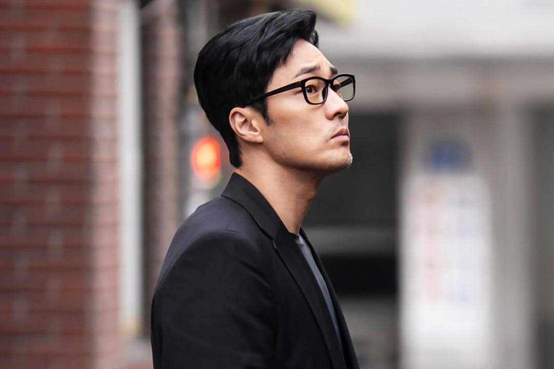 Seorang pria berjas hitam dan berkacamata berdiri di luar ruangan dengan ekspresi serius dalam adegan drama Agent Kim Reactivated.
