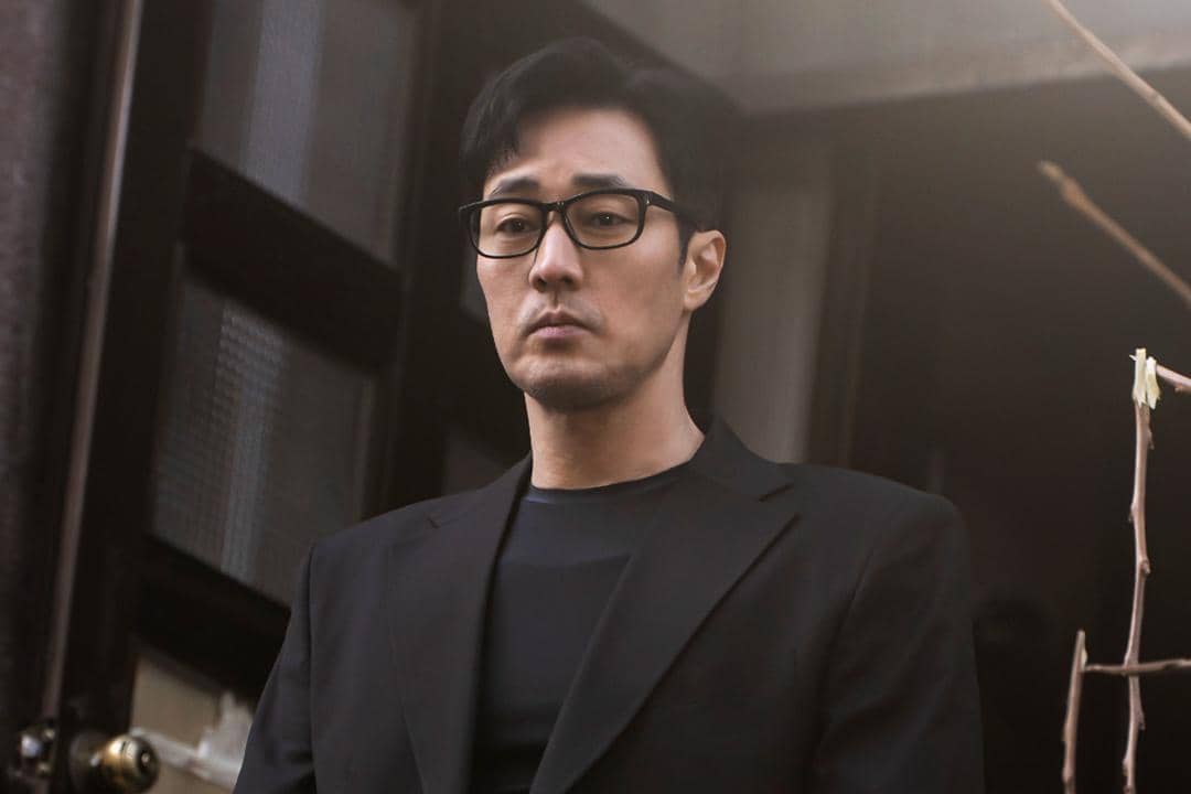 Seorang pria berpenampilan serius mengenakan jas hitam dan kacamata berdiri di depan pintu dalam cuplikan drama Agent Kim Reactivated.