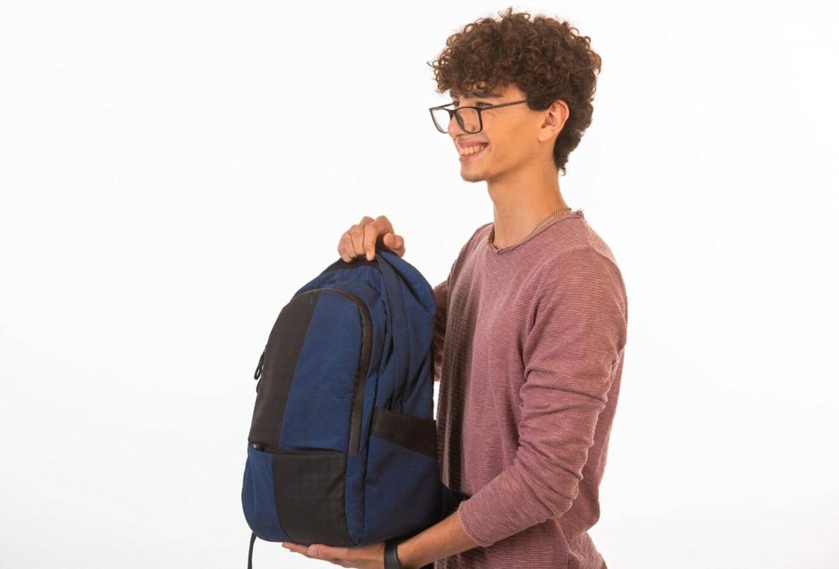 Seorang pria muda berkacamata mengenakan kaus merah muda memegang ransel biru di depan tubuhnya dengan latar belakang putih.