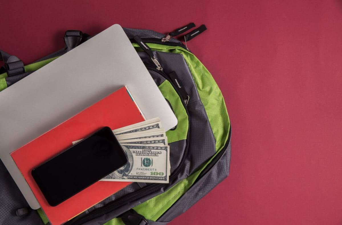 Tumpukan uang dolar, ponsel, buku catatan, dan laptop di atas ransel abu-abu hijau dengan latar belakang merah marun.