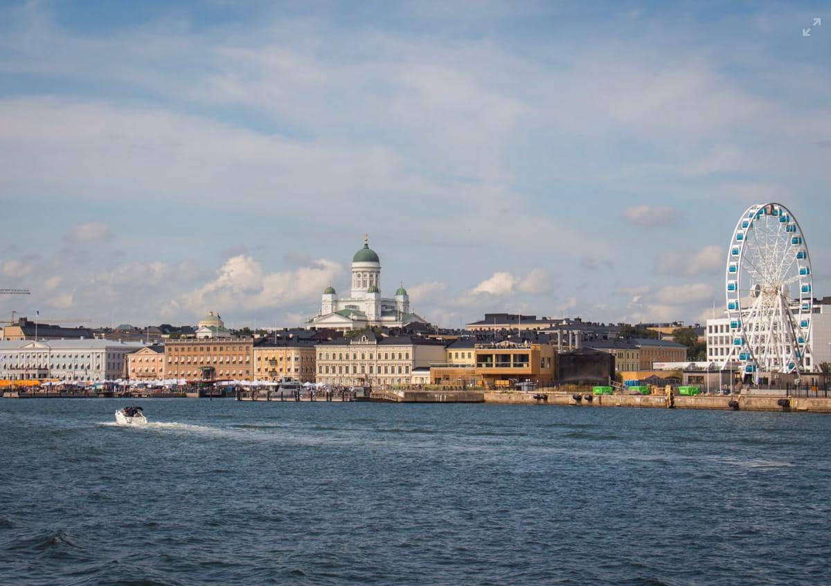 Pemandangan kota Helsinki di tepi laut dengan katedral berkubah hijau dan bianglala besar di bawah langit cerah Finlandia.