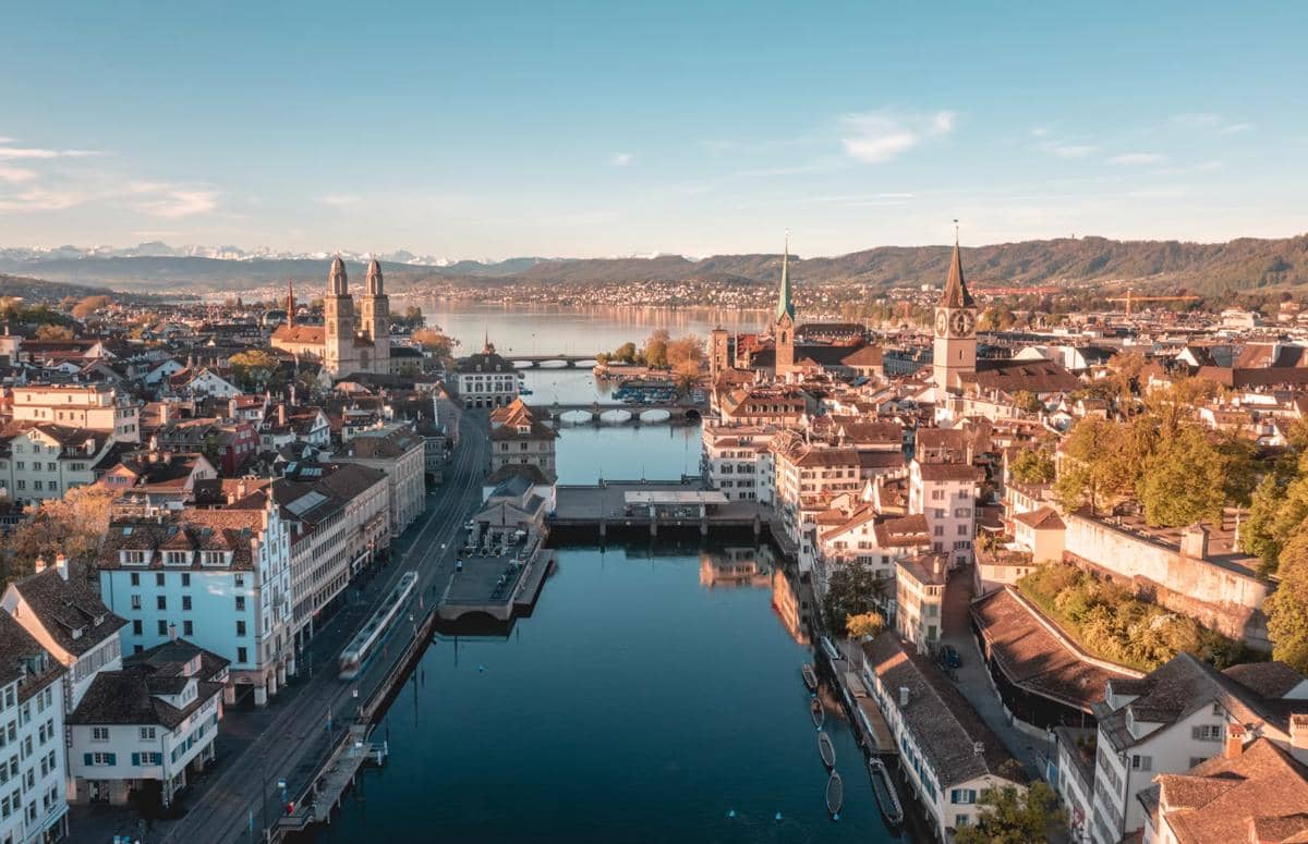 Pemandangan udara kota Zurich di Swiss dengan sungai, bangunan bersejarah, dan pegunungan di kejauhan di bawah langit cerah.