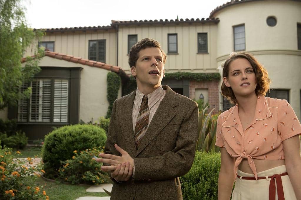 Dua karakter berdiri di taman depan rumah bergaya klasik dalam adegan film Cafe Society, mengenakan busana era 1930-an.