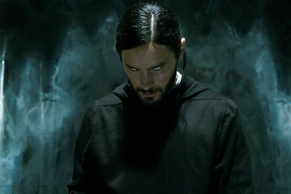 Seorang pria berwajah pucat dengan mata putih menatap tajam ke bawah di ruangan gelap bersuasana misterius dari film Morbius.