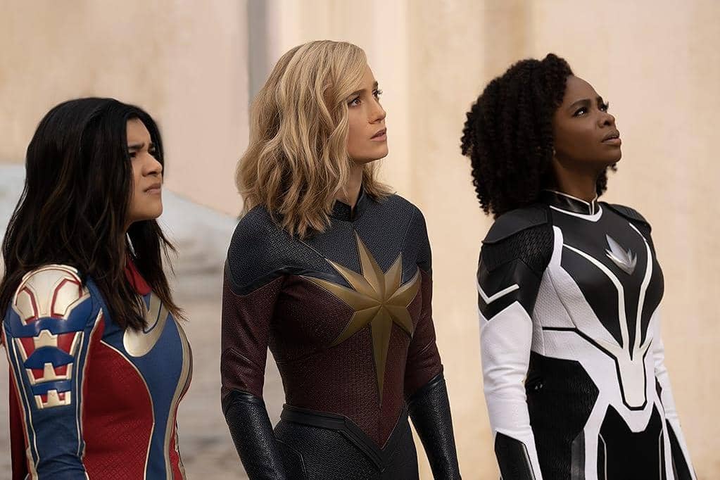 Tiga pahlawan perempuan mengenakan kostum superhero berwarna merah, biru, dan hitam putih berdiri berdampingan menatap ke arah atas.