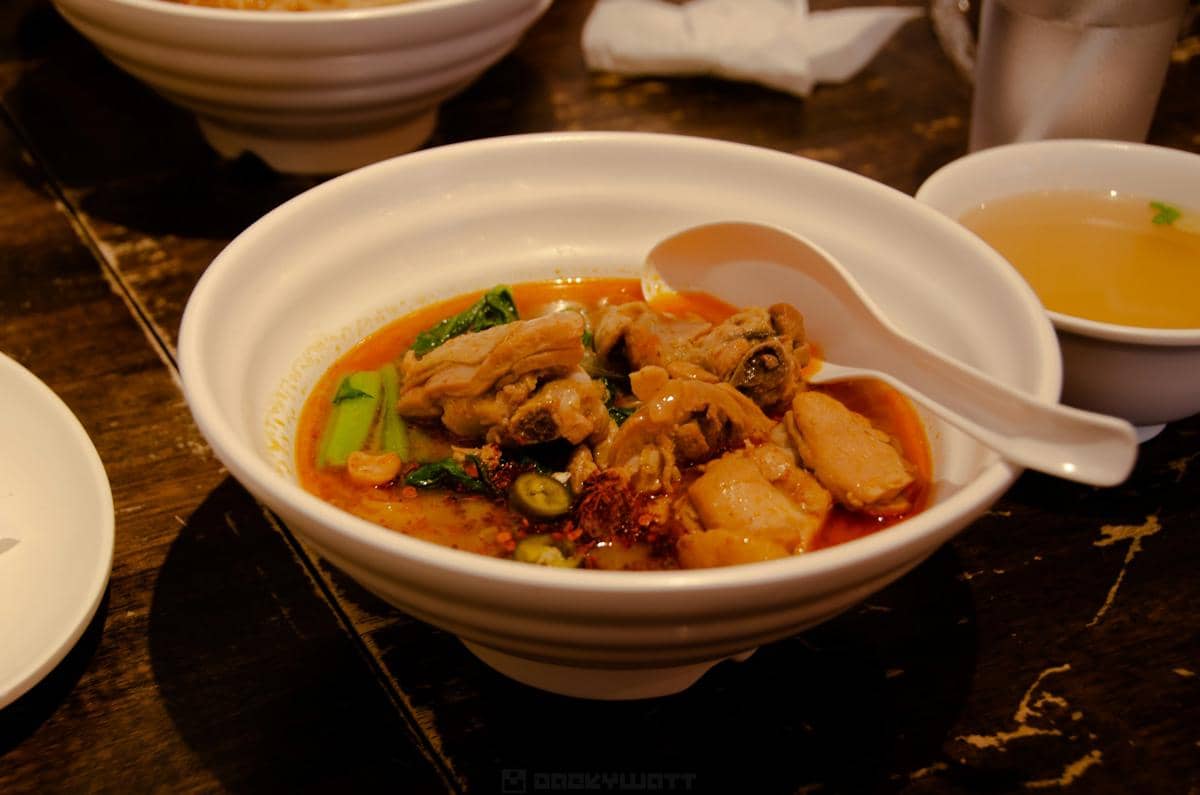 Semangkuk hidangan ayam velvet berkuah merah dengan potongan ayam, sayuran hijau, dan bumbu pedas disajikan di atas meja kayu.