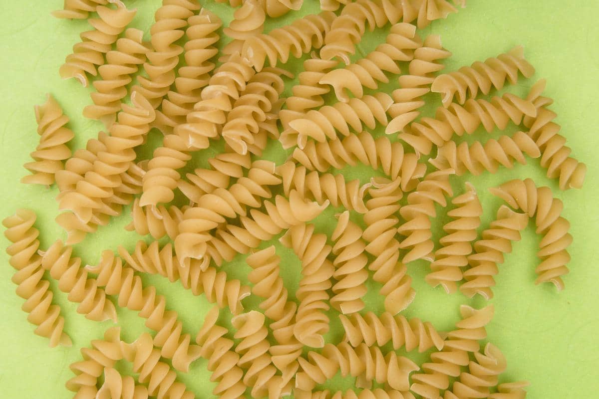 Beberapa potong pasta fusilli mentah berwarna kuning muda tersusun di atas permukaan hijau terang untuk bahan salad pasta.