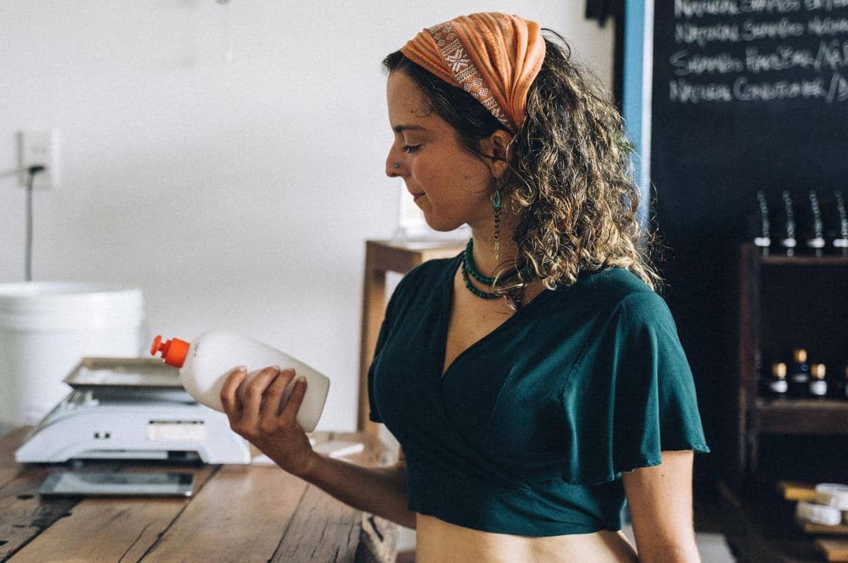 Seorang wanita mengenakan bandana oranye dan atasan hijau sedang memegang botol plastik putih di dalam ruangan dengan meja kayu.