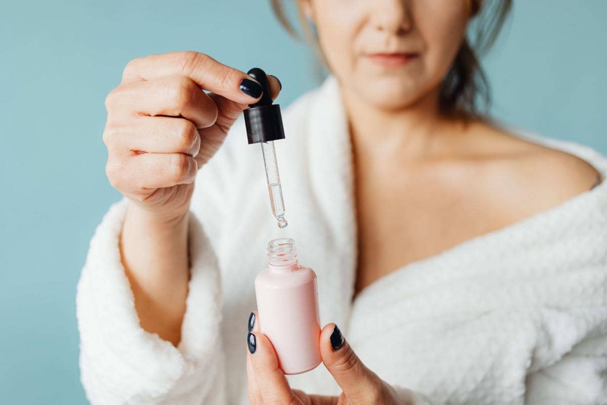 Seorang wanita mengenakan jubah mandi putih memegang botol serum berwarna pink dan pipet tetes di tangan dengan latar biru lembut.