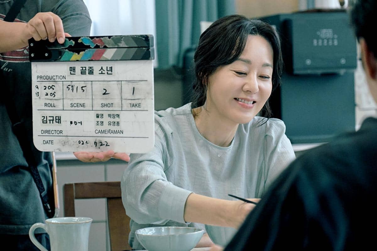 Seorang aktris tersenyum saat syuting adegan di meja makan dengan papan klak film bertuliskan Notes from the Last Row dipegang kru.