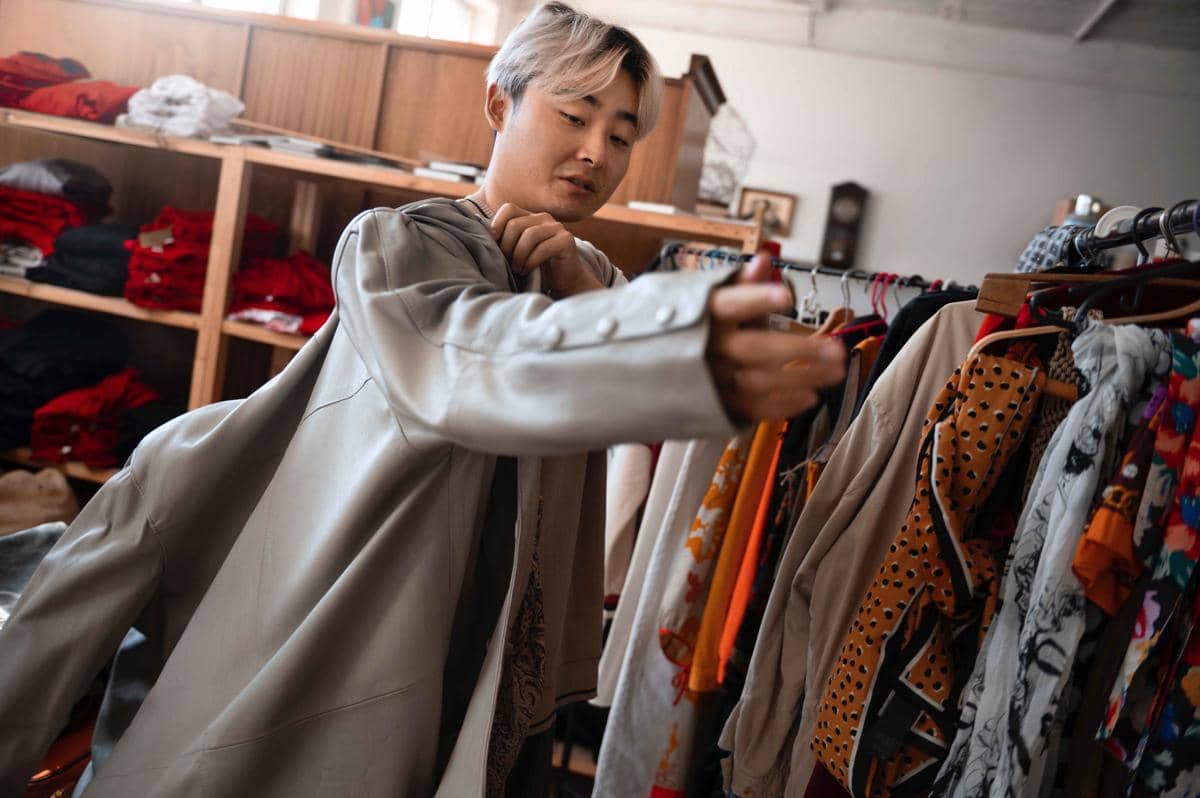 Seorang pria mencoba jaket sambil memilih pakaian di toko barang bekas dengan rak penuh baju berwarna-warni di sekitarnya.