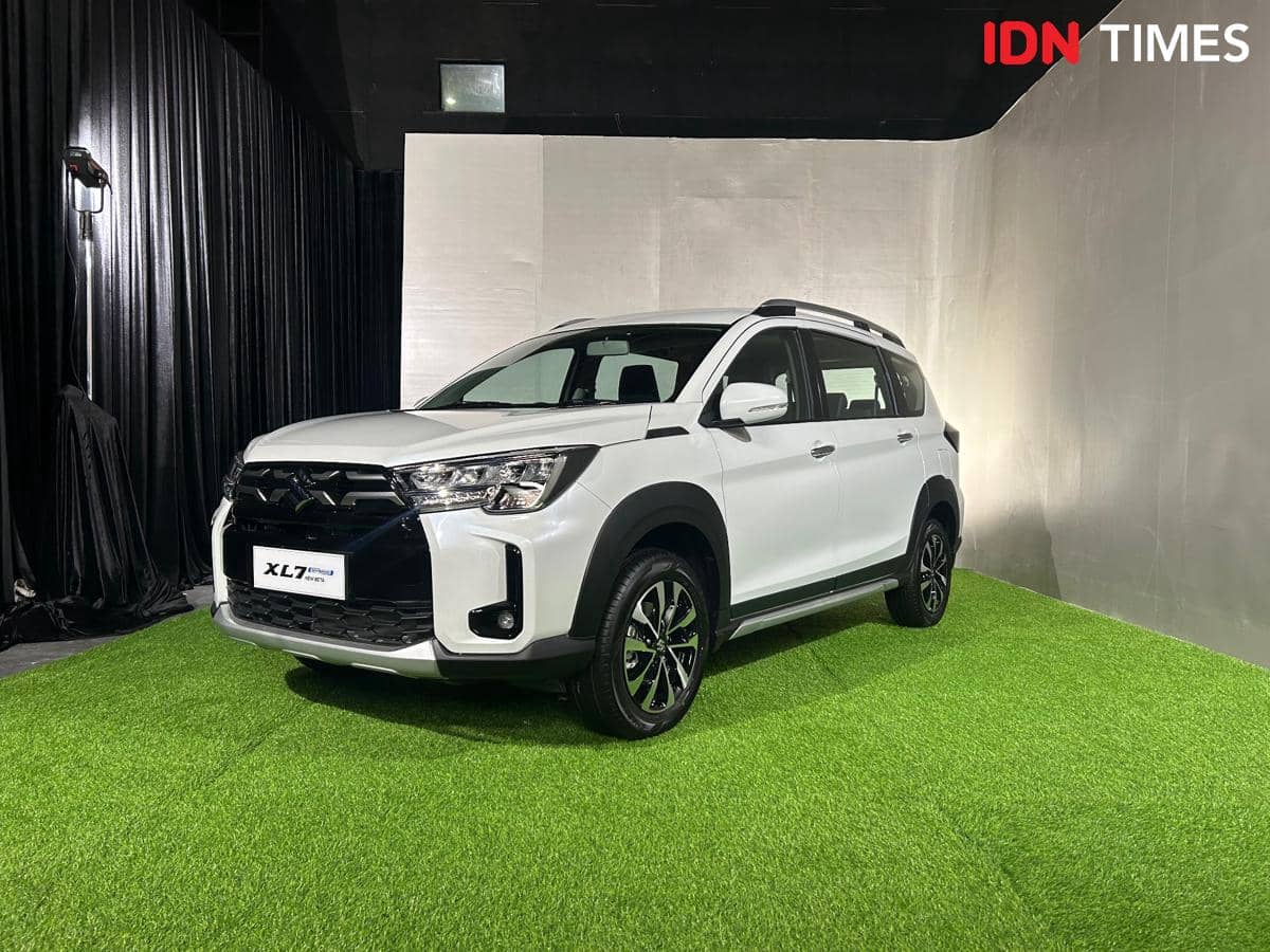 Mobil Suzuki New XL7 Hybrid berwarna putih dipamerkan di dalam ruangan dengan lantai rumput sintetis dan latar hitam abu-abu.