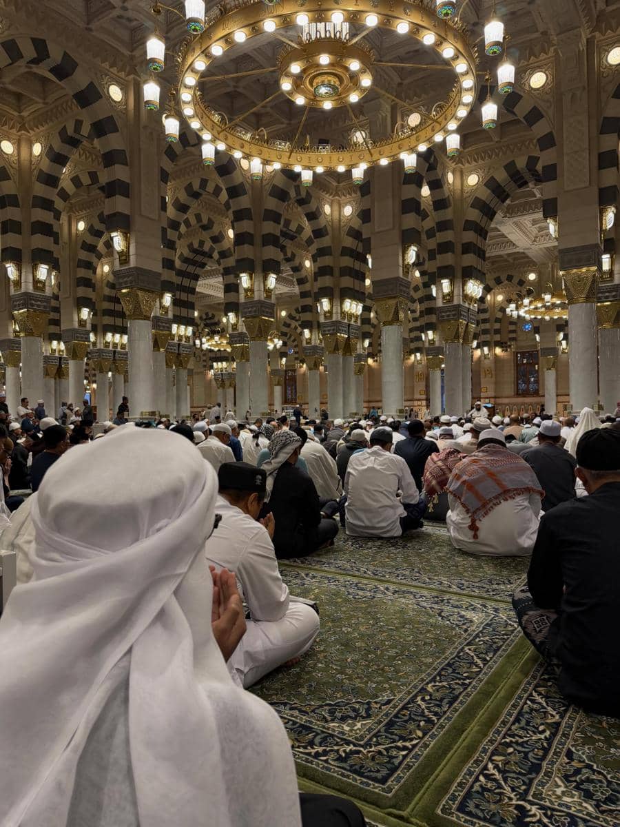 Jemaah mengenakan pakaian ihram putih sedang berdoa di dalam masjid megah dengan lampu gantung besar dan pilar berornamen emas.