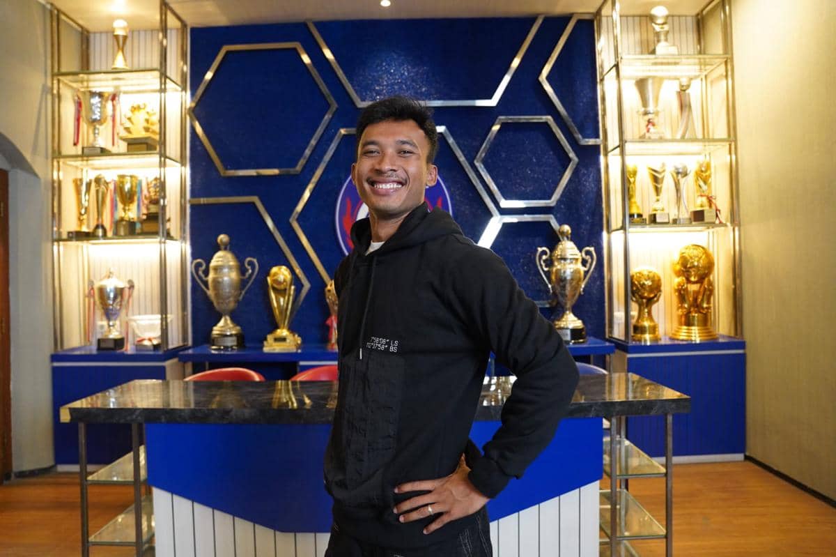 Seorang pria mengenakan hoodie hitam berpose sambil tersenyum di depan deretan trofi dan latar berwarna biru bertema Arema FC.