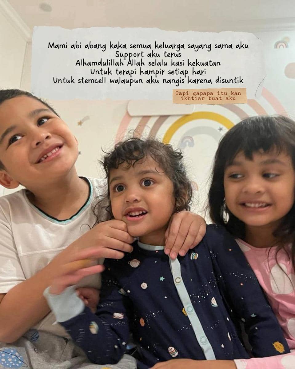 Tiga anak kecil duduk bersama di dalam ruangan dengan dinding bergambar pelangi, tersenyum dan saling berpelukan dengan hangat.
