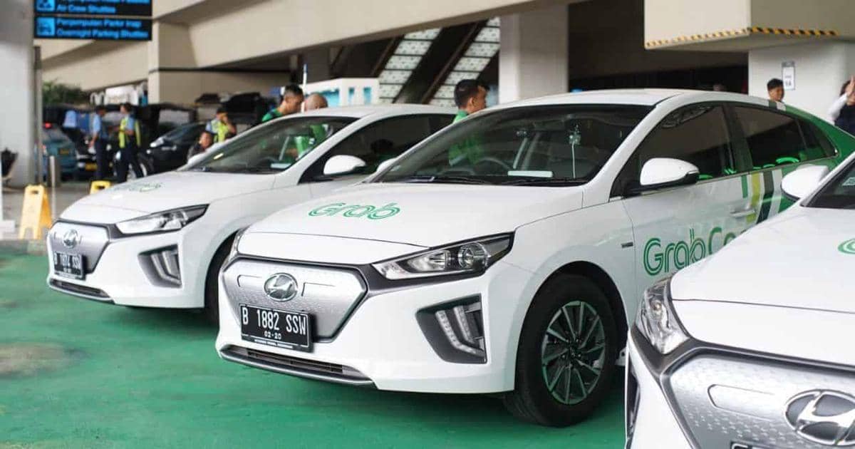 Beberapa mobil GrabCar berwarna putih berlogo Grab terparkir berjejer di area parkir dengan pengemudi berdiri di dekatnya.