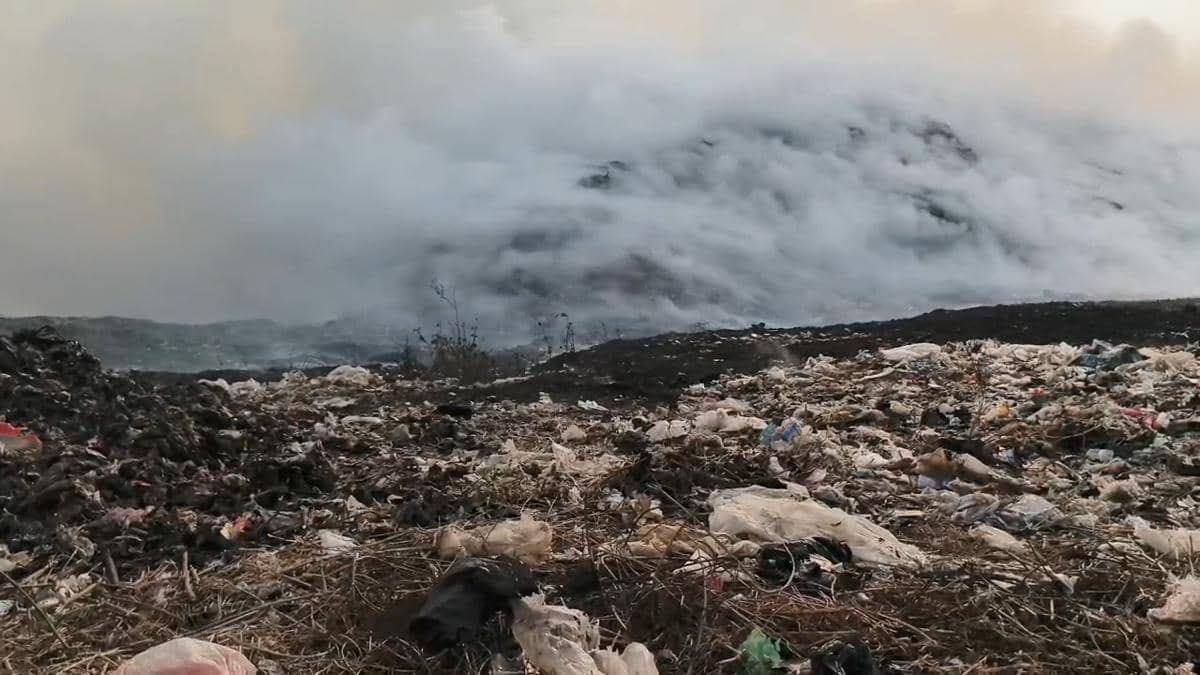 Asap tebal mengepul dari tumpukan sampah yang terbakar di Tempat Pembuangan Akhir Jatiwaringin, dengan sisa-sisa sampah berserakan di sekitarnya.