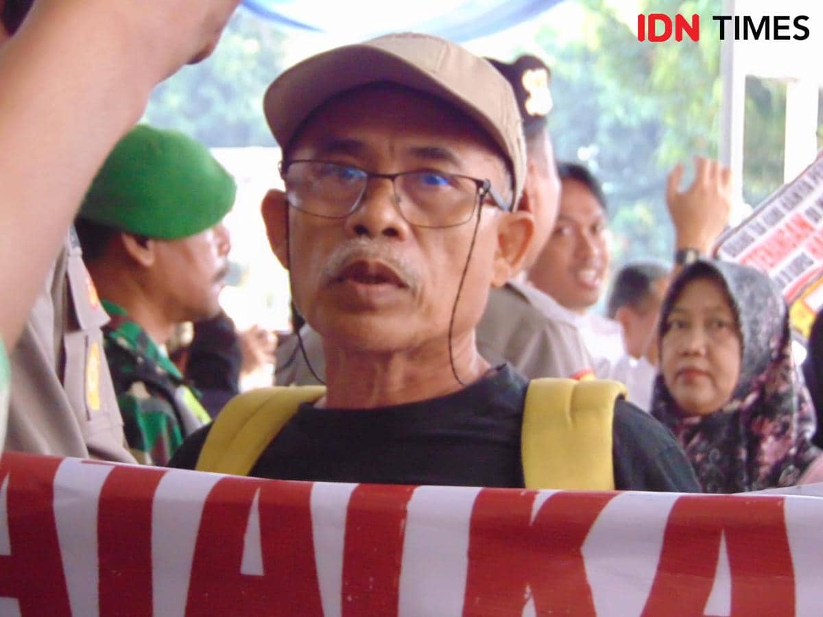 Seorang pensiunan mengenakan topi dan kacamata memegang spanduk saat aksi protes di depan Bank Mandiri Taspen Purwokerto.