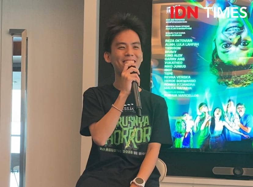 Seorang pria mengenakan kaus hitam bertuliskan 'Harusnya Horor' sedang berbicara dengan mikrofon di depan poster film di SCTV Tower.