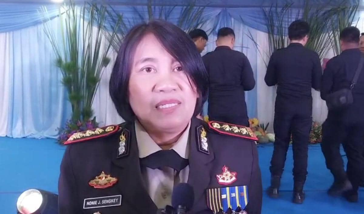 Seorang perwira polisi berpakaian dinas lengkap dengan tanda pangkat dan medali berbicara di sebuah acara resmi dengan latar biru.