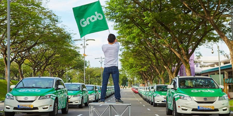 Seorang pria berdiri di tengah jalan sambil mengibarkan bendera Grab di depan deretan mobil Grab berwarna hijau putih di bawah pepohonan rindang.