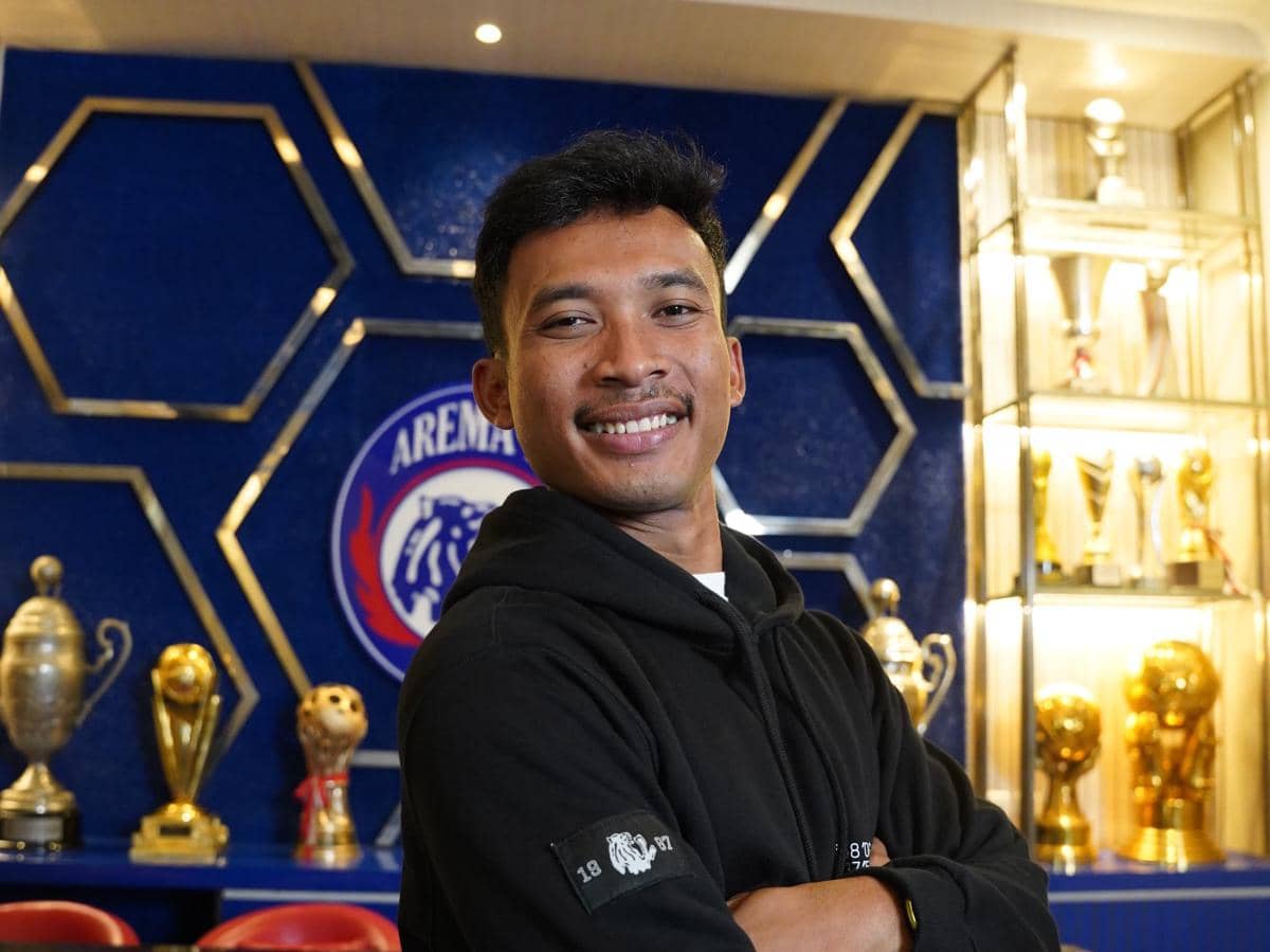 Seorang pria mengenakan hoodie hitam berpose tersenyum di depan logo Arema FC dengan deretan trofi emas di latar belakang.