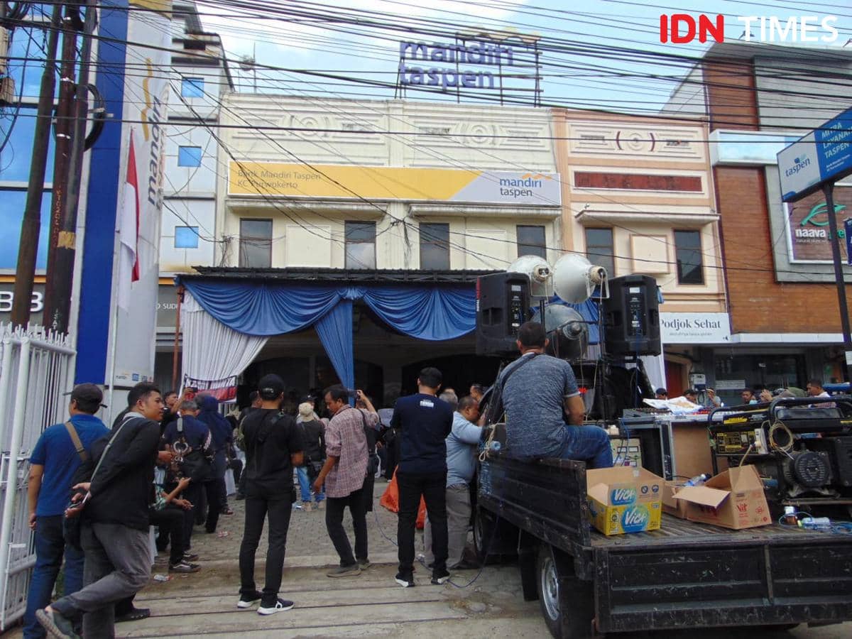 Sejumlah orang berkumpul di depan kantor Bank Mandiri Taspen Purwokerto dengan peralatan suara dan kendaraan di area parkir.